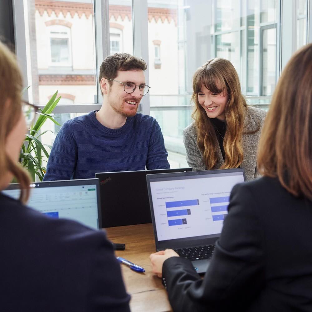 Colleagues discuss and work together at a table on their laptops.