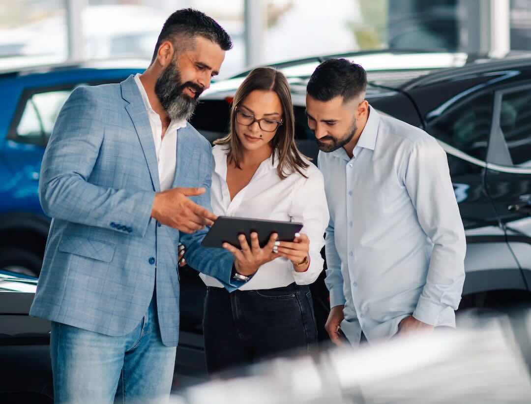 Two customers and a car dealer look at a tablet together in the showroom.