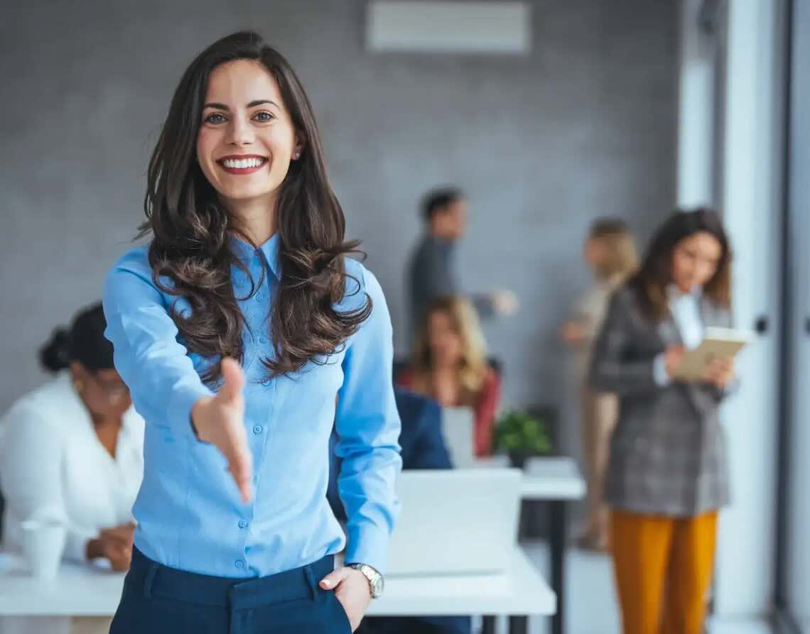 Woman extends her hand in greeting in an office full of employees.
