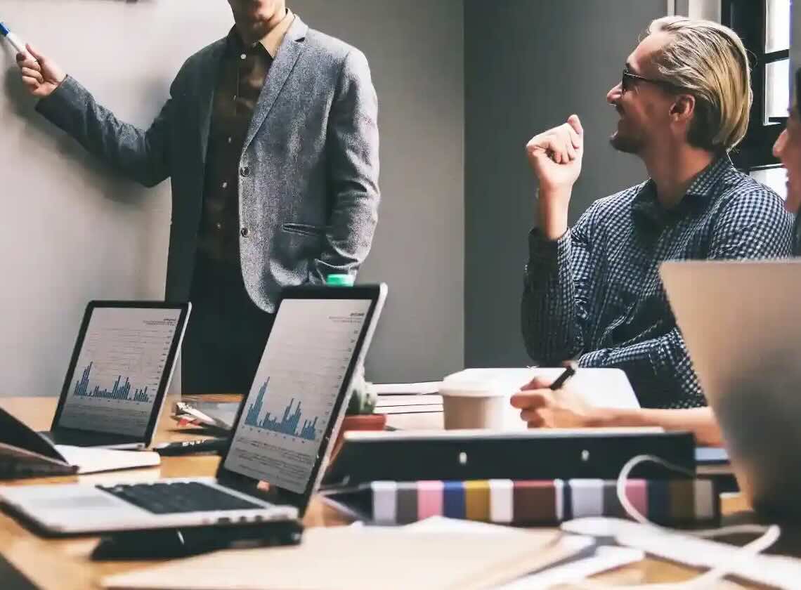People with open laptops sit together in a meeting room and discuss something on the whiteboard.