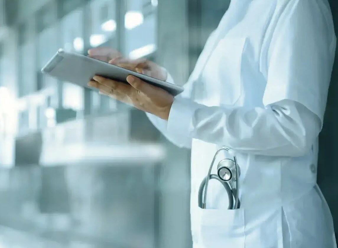 A doctor in scrubs and with a stethoscope in his coat pocket stands in the hospital corridor and checks information on their tablet.