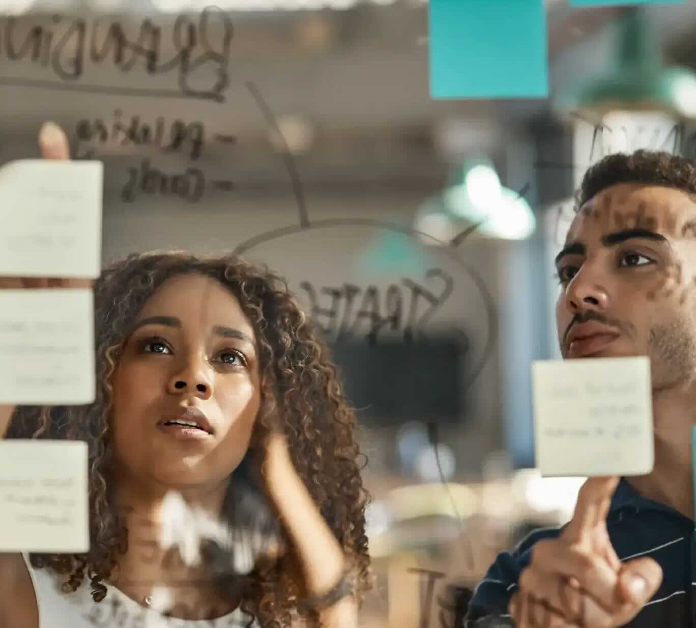 Two people collaborate together standing at a glass whiteboard with post-its and text.