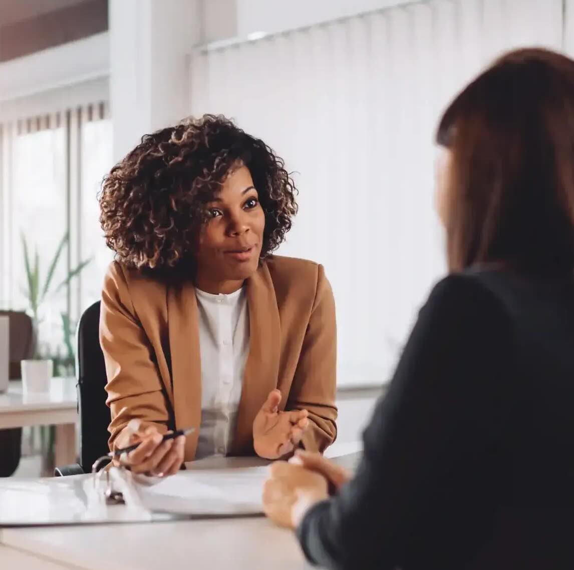 Two women are sitting opposite each other in an office room. One woman shows the other something in a folder.