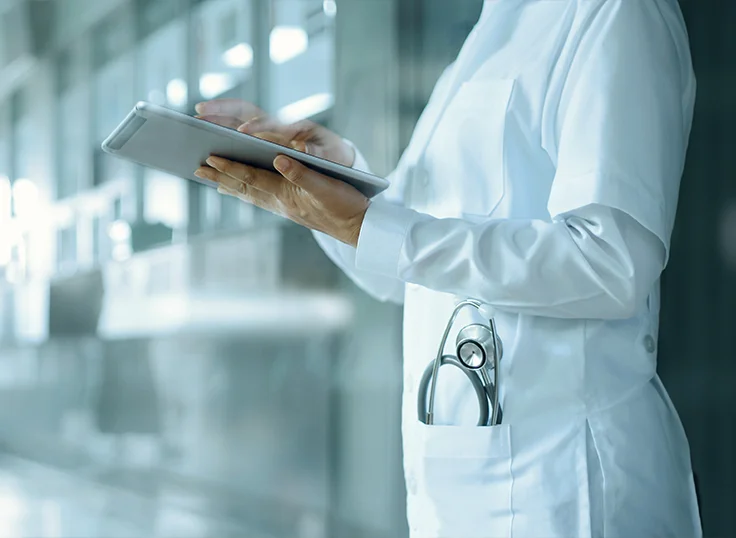 A doctor in scrubs and with a stethoscope in his coat pocket stands in the hospital corridor and checks information on their tablet.