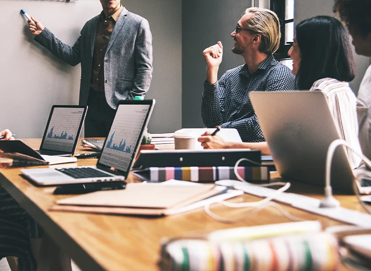 People with open laptops sit together in a meeting room and discuss something on the whiteboard.