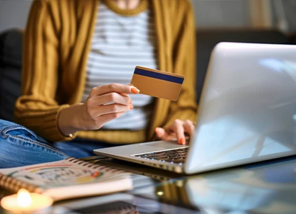 A woman sits in her living room on the couch in front of her laptop and orders something with her credit card.